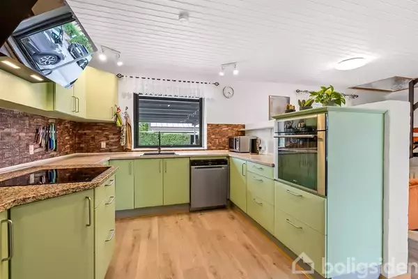 Et spisebord med fire stole står i et lyst rum med trægulv. En stor glasrude viser en frodig have udenfor. En kommode ses mod en væg.