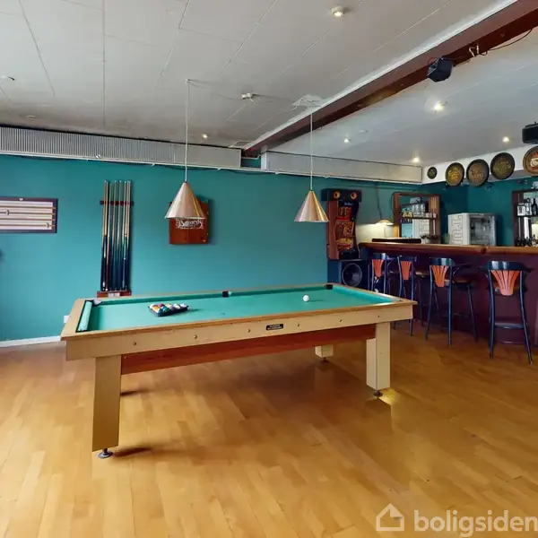 Poolbord i centrum med kuglerne klar til spil. Omkring bordet er der en bar med høje stole og hylder fyldt med glas, belyst af lamper. Væggene har spildekor.