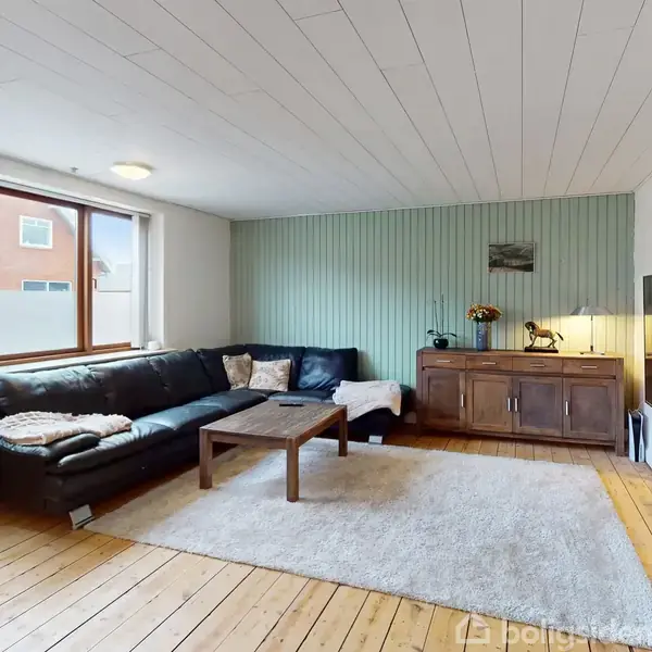 En sort hjørnesofa står ved et vindue i en stue med lyse trægulve. Et træbord foran sofaen, et fladskærms-TV på væggen, og et skab med dekorationer ved siden af.