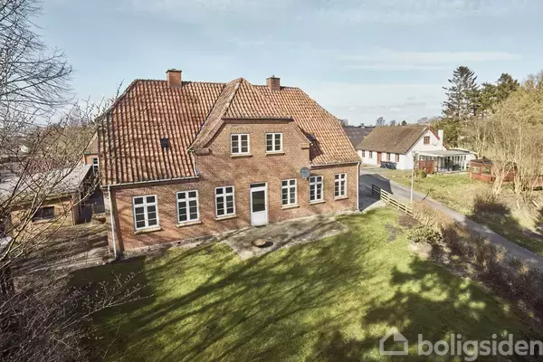 En rød murstensvilla med rødt tegltag står stille i et grønt haveområde ved en vej. Bagved ses flere huse og træer i en landskabelig, landlig setting.