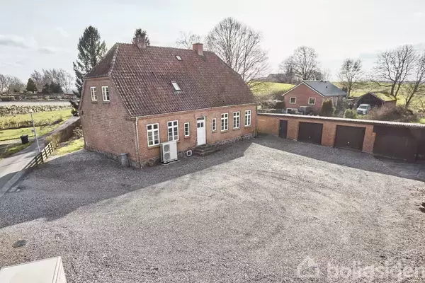 Et rødstenshus med skråt tag står på en grusbelagt plads. Bagved er garager og en have med træer. Huset er omgivet af en landevej og åbne marker.