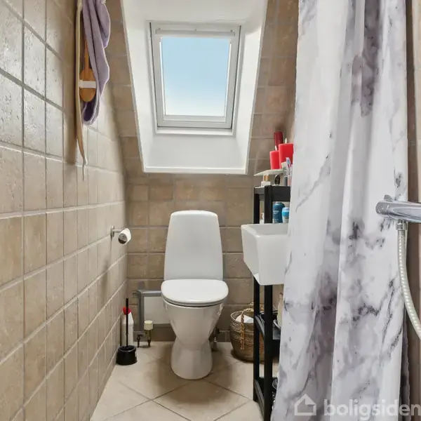 Toilet står i et lille badeværelse med flisevægge og skråvindue. En bruser er til højre bag et forhæng, og en lille håndvask med hylde står ved siden af toilettet.