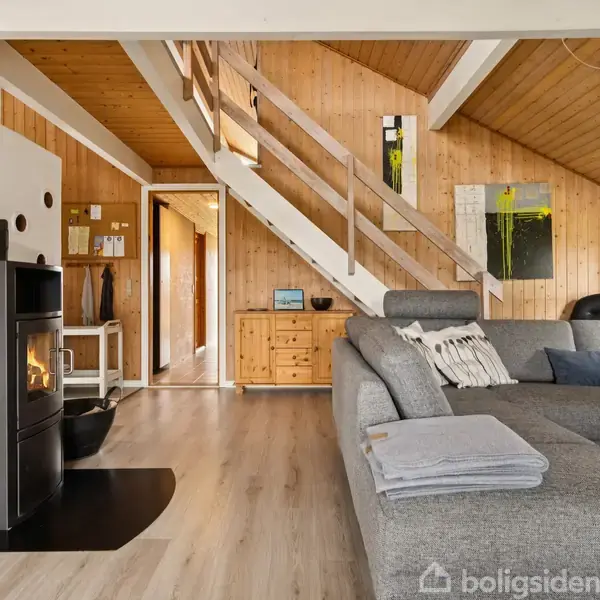 Pejsen brænder i en stue med grå sofa, trævægge, og en trappe leder til en hems. Rummet er lyst og indbydende med moderne indretning.