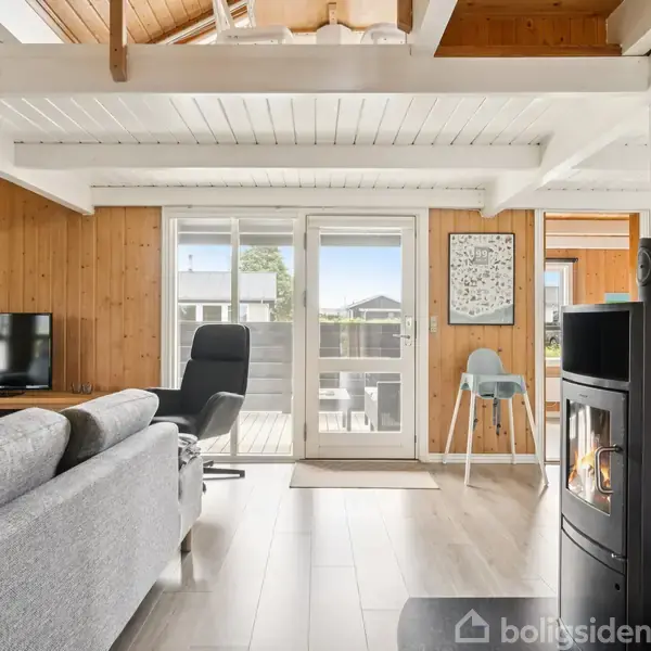 Pejs i stue tændt, giver varm atmosfære. Grå sofa og lænestol vender mod TV i rum med trævægge og stort vindue mod terrasse. Lys moderne indretning.