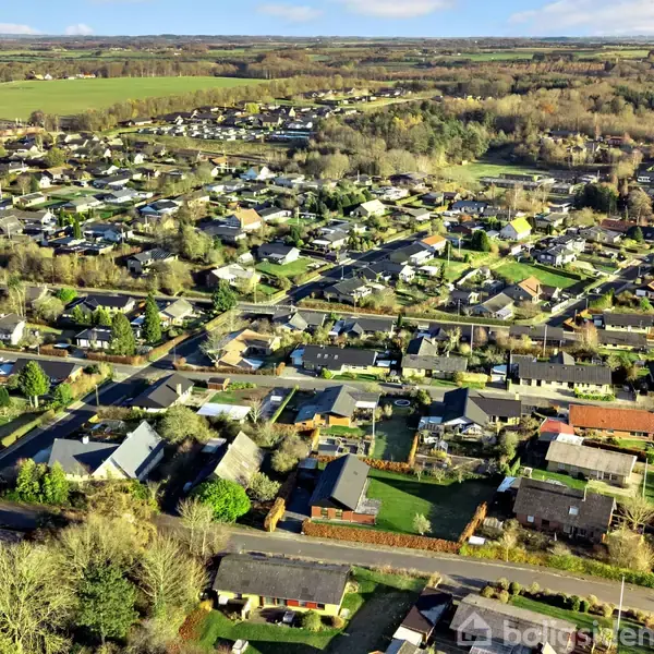 Beboelseskvarteret strækker sig med huse og veje. Omgivet af grønne marker og skove, vises et typisk dansk forstadsområde med spredte træer og velholdte haver.