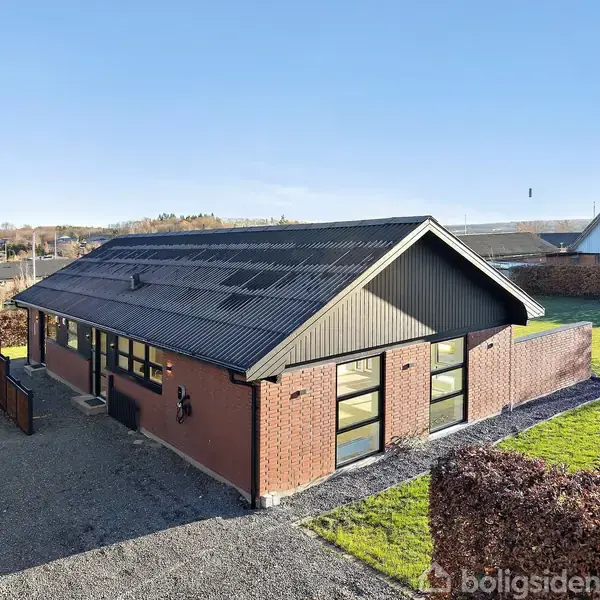 En rødstensbungalow med sort tag står omkranset af en velholdt have med græsplæne og hække. Beliggende i et beboelseskvarter under en blå himmel.