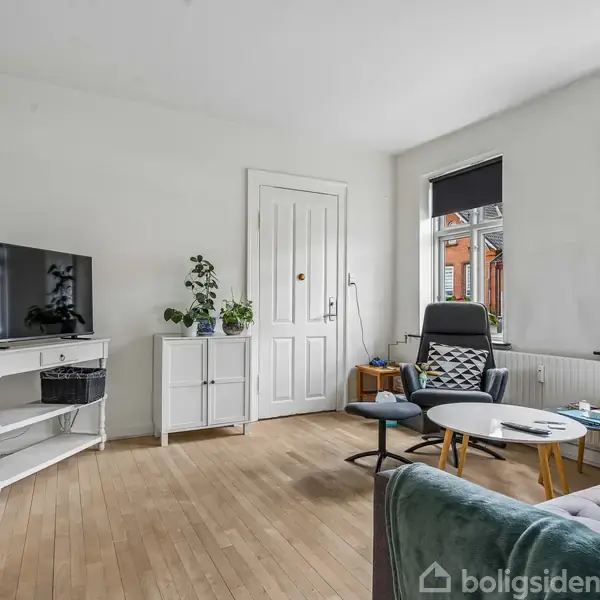 Stue med fladskærms-tv på hvidt bord; sofa, lavt rundt bord med pynt og plantestander ved vindue; lyst trægulv og to store vinduer med udsigt til røde murstenshuse.