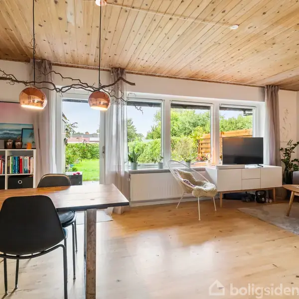 Stue med træloft og trægulv, spisebord med sorte stole, hængende lamper, sofa foran kunst på væggen, stort vindue med udsigt til have, TV i hjørnet.