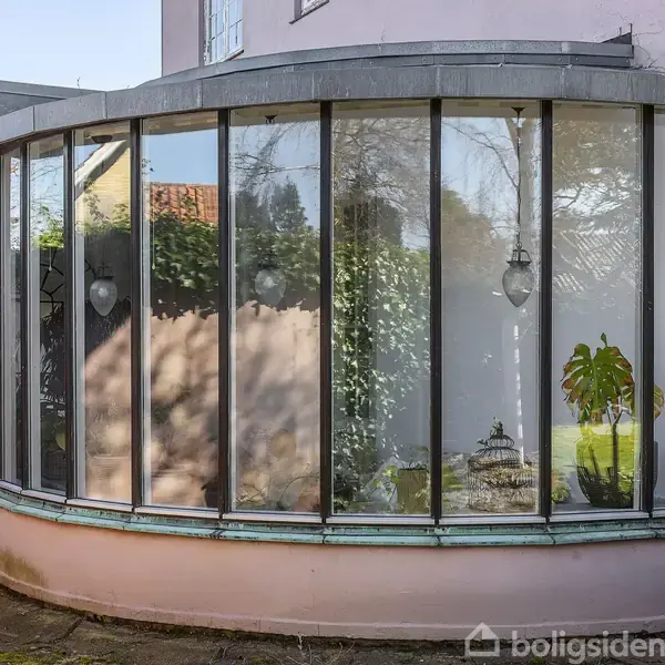 Et buet vinduesparti viser refleksioner af en have med træer og buske. Indenfor står en grøn plante nær vinduet. Boligens rosa facade er synlig i baggrunden.