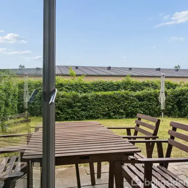 Et træbord med tilhørende stole står på en terrasse. Udsigt gennem en glasdør viser en have omkranset af en hæk under en blå himmel med få skyer.