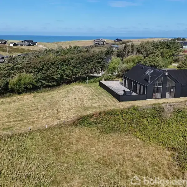Et sort sommerhus står stille i en grøn, bakket landskab med havudsigt i baggrunden. Omgivet af græs og spredte træer, er der flere huse i nærheden.