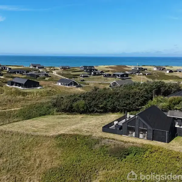Et sort sommerhus står stille på en græseng, omgivet af flere lignende huse på kuperede bakker nær kysten. I baggrunden ses havet under en klar blå himmel.