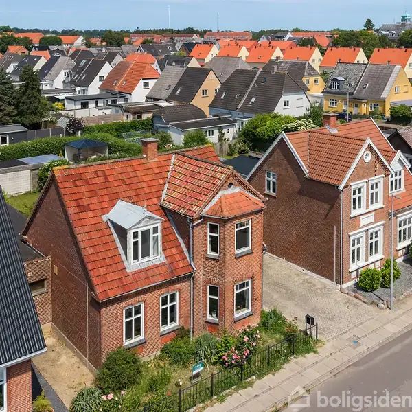 Røde murstenshuse står på række med forskelligt farvede tage, omgivet af frodige haver og en asfalteret vej, i en velordnet boligkvarter med flere huse i baggrunden.