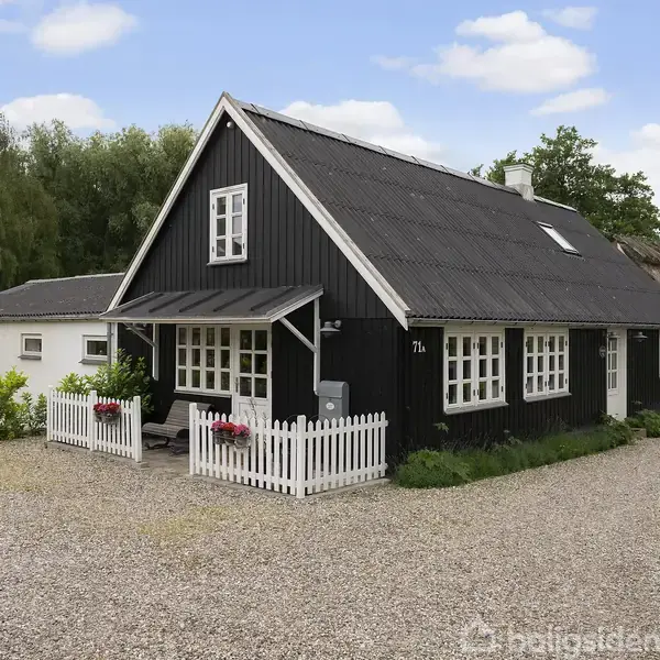 Et sort træhus med hvide vinduer og en lille terrasse med blomsterkrukker står på en grusvej. I baggrunden er der grønne træer og andre huse.