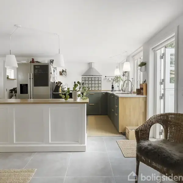 Køkken med hvid køkkenø og træbordplade, omgivet af planter og stålapparater. Lyse fliser på gulvet, to hængende lamper, åbent til spisestue til venstre, kurvestol til højre.
