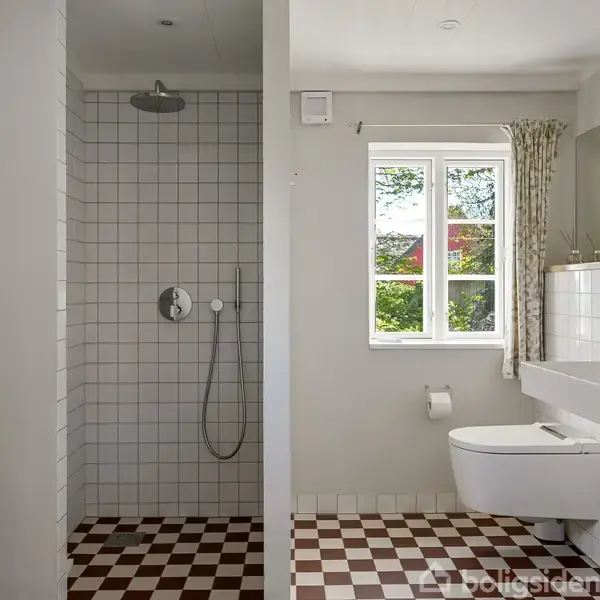 En bruser i et badeværelse med hvide fliser står til venstre, mens en håndvask og toilet er installeret ved et vindue til højre. Skakternet gulv afslutter scenen.