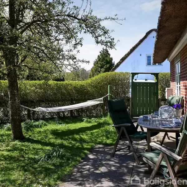 Hængekøje hænger mellem træ og hæk i grøn have. Rundt bord med stole står ved siden af murstenshus med stråtag. Dør fører til nabogrund. Ingen tekst til stede.