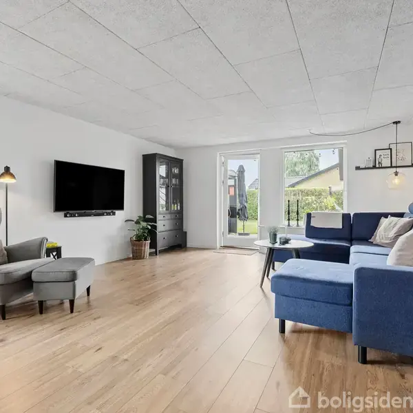 Stue med blå hjørnesofa og lænestol, fladskærms-TV på væggen, trælaminatgulv og sofabord. Store vinduer med udsigt til have giver lys til rummet.
