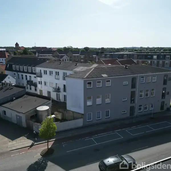 Bygning med fladt tag og hvide og grå facader står stille i et bymiljø. Omkring er parkerede biler og træer langs vejen. Baggrunden viser flere huse og en kirke.