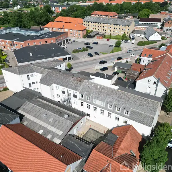 Bygning med hvid facade ses fra oven, beliggende i et byområde omgivet af andre bygninger med røde tage. Biler er parkeret i nærheden, omkranset af grønne træer.