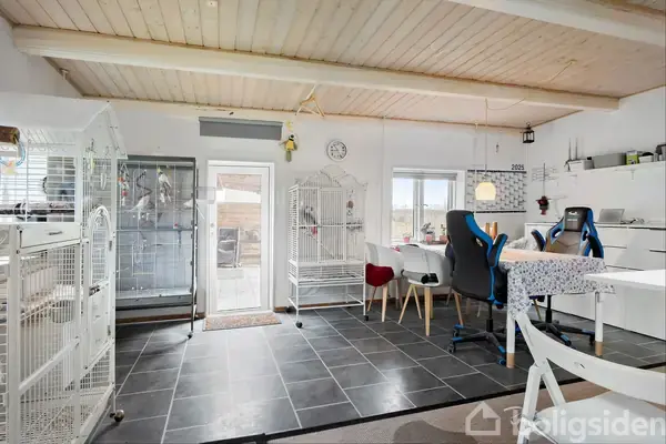 Fuglebure står stille i et lyst, flisebelagt rum med hvidt loft. Der er et bord med stole, en vægklokke og dekorative elementer. Vinduet og døren lader naturligt lys komme ind.