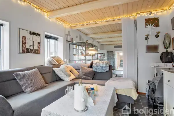 Grå hjørnesofa fyldt med puder står i en stue med træloft. Små lyskæder dekorerer rummet, hvor billeder hænger på væggen, og et spisebord ses i baggrunden.
