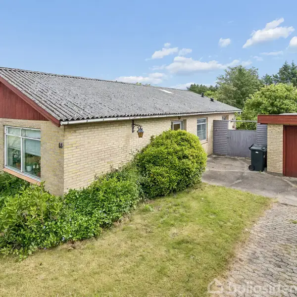 Et enfamiliehus med gul murstensfacade og gråt tag, omgivet af en grøn have. Terrassen fører til en tilhørende garage med rød port. Beliggende i et skovområde.