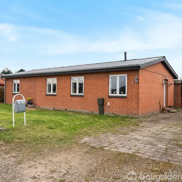 En rød murstensvilla står på en grøn græsplæne med en indkørsel af fliser. Huset har flere vinduer, og en grill står til højre for huset tæt på garagen.