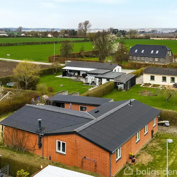 En rød murstensbygning med sort tag står stille omgivet af grønne marker og naboer, inklusive en større moderne bygning. Der er indkørsler og haver i området.
