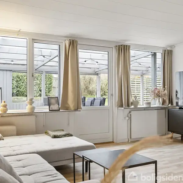 En lys beige sofa står i ro mod en cremefarvet væg i en hyggelig stue, flankeret af et sort sofabord og en grå skænk. En lampe hænger i hjørnet.