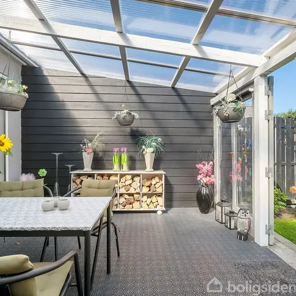 Et hyggeligt, overdækket terrasseområde med stole omkring et bord. Blomsterdekorationer hænger fra loftet. Et skab mod en trævæg indeholder brænde. Udsigt til have gennem glasdøre.
