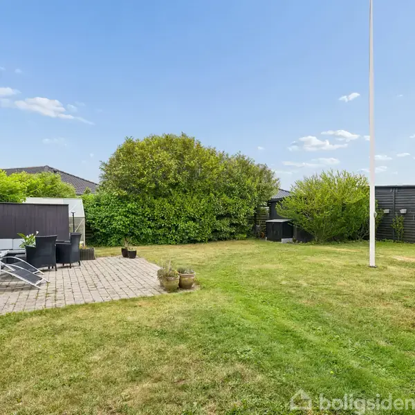 En terrasse med havemøbler står på en flisebelagt plads, omgivet af en græsplæne og grønne buske. I baggrunden ses et lille skur og en flagstang under en klar blå himmel.