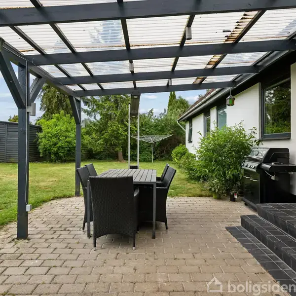 En overdækket terrasse har sorte stole og et bord på et flisebelagt område. Til højre står en grill ved siden af husets indgang. Græsplæne og buske omkranser området.