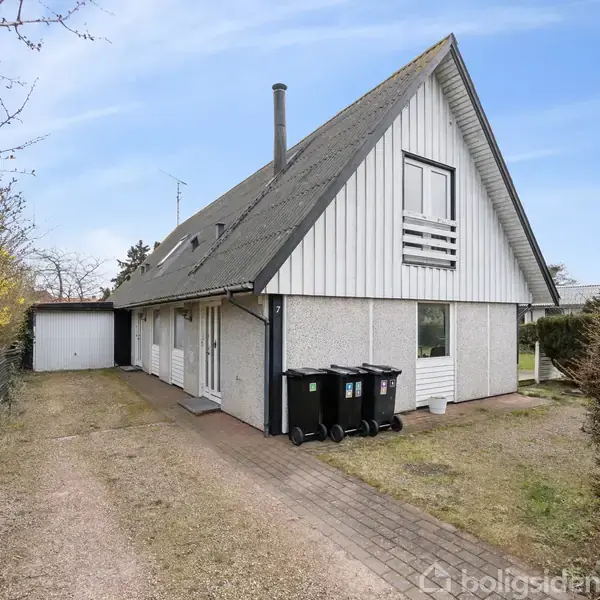 Et hvidt, trekantet hus med skråt tag står ved en grusvej. Tre skraldespande er placeret ved siden af huset. En garage ses i baggrunden mellem grene og hegn.