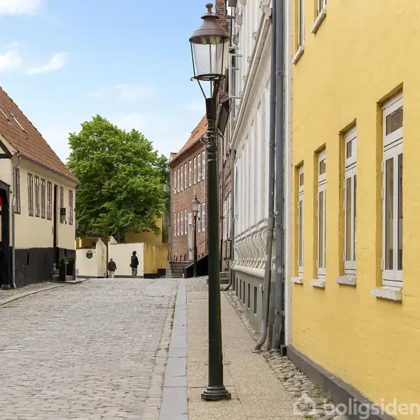 En stille brostensgade omgivet af gule og røde murstensbygninger, med gadelamper langs fortovet. En person går væk fra kameraet mod et grønt træ i enden af gaden.