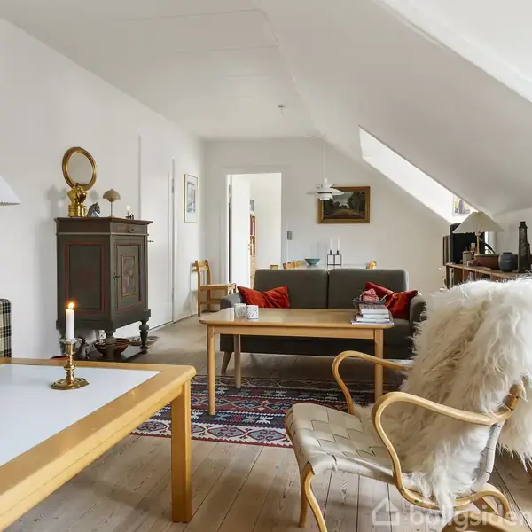 Stue med møbler, herunder et sofabord med tændt stearinlys og en stol med hvidt lammeskind. Rummet inkluderer en sofa, tæppe og en reol, med skrånende lofter.