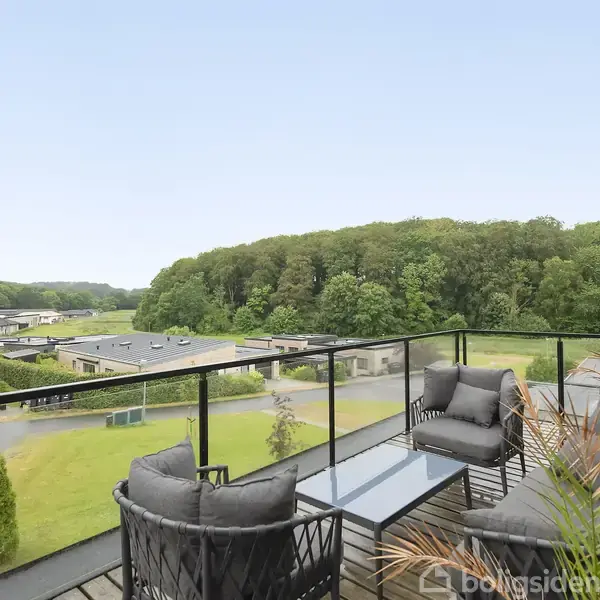 En moderne udendørs terrasse med polstrede møbler og glasbord står stille, omgivet af grønne trækroner og lave bygninger i baggrunden under en klar, blå himmel.