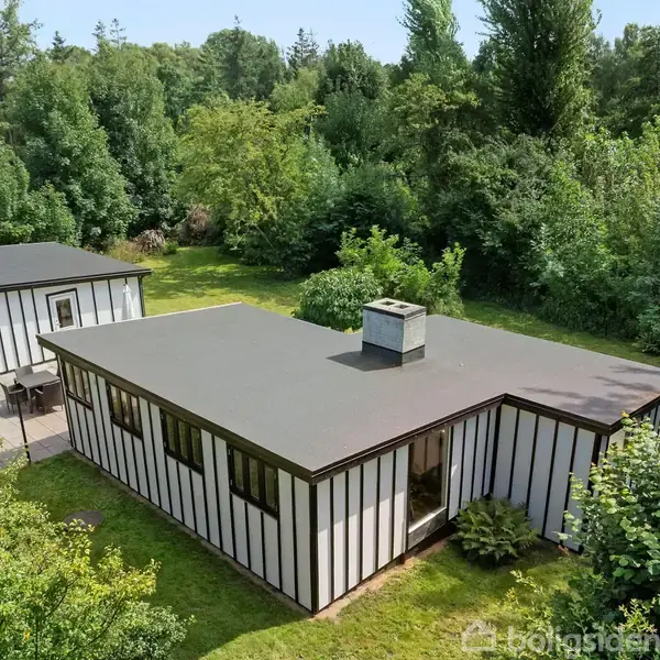 Et moderne, minimalistisk sommerhus med fladt tag og store vinduer står midt i en frodig, grøn have. Et fritstående skur ved siden af huset er omgivet af træer og buske.