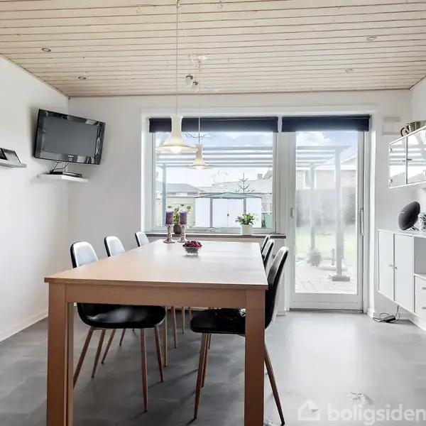 Langt spisebord med seks stole står i spisestue med lyst trægulv og hvide vægge. Et vægbeslag bærer et fjernsyn, og store vinduer giver udsigt til haven. Hylder med pynt på væggen.