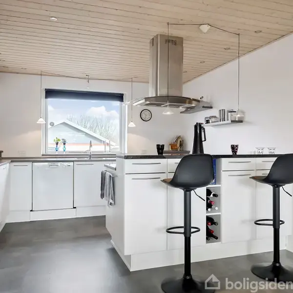 Et moderne køkken med en hvid køkkenø, to sorte barstole og en kogeplade med emhætte. Et stort vindue giver udsigt til et sneklædt landskab.