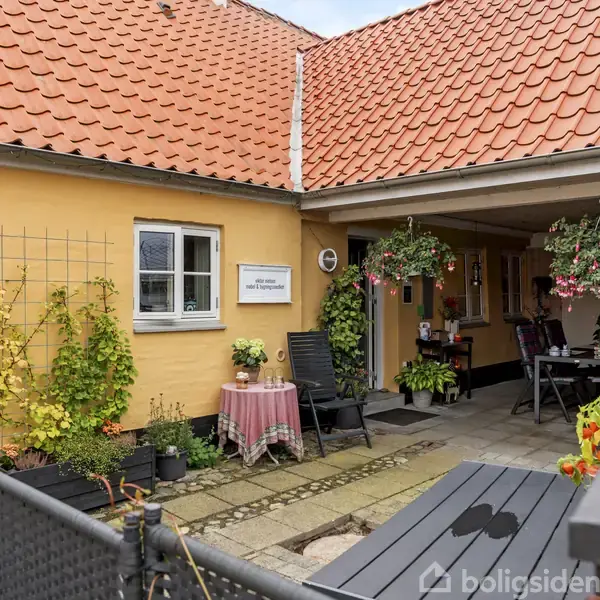 Gul væg med klatreplanter og et skilt siger "Velkommen til Solgården." Til venstre står et lille bord med dug. I baggrunden er en overdækket terrasse med borde og stole.