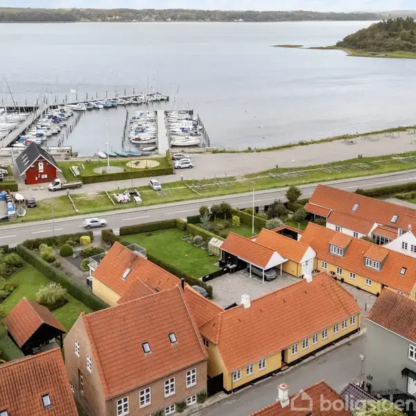 Røde tage ligger tæt sammen i en lille by ved havet, hvor en marina med mange både ses ved kysten i det fjerne. En vej adskiller husene og marinaen.