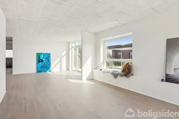 Et lyst værelse med trægulv og hvidmalede vægge. En blå plakat står på gulvet. Sollys skinner ind gennem store vinduer. En pude og tæppe ligger på en bred vindueskarm.