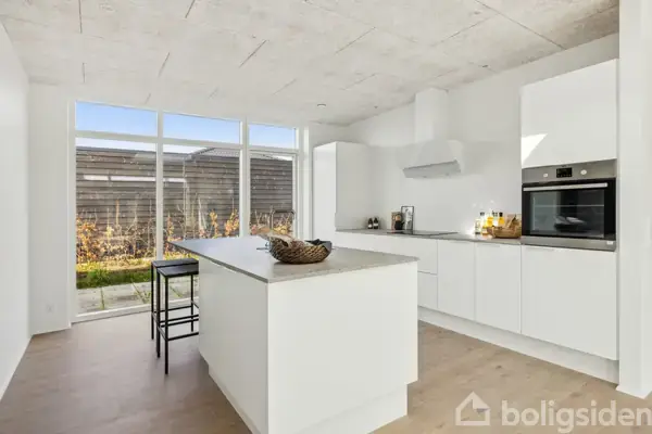 En moderne køkkenø i aktion med en kurv på toppen, beliggende i et lyst køkken. Rummet har store vinduer, hvidt interiør og trægulve med udsigt til en have.