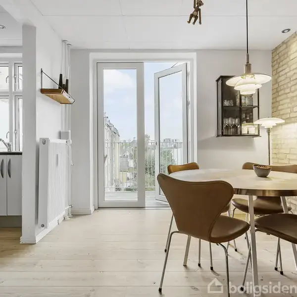 Rundt spisebord med fire stole står i et lyst, moderne rum. Dobbeltglasdør åbner mod en balkon. Rummet har en murstensvæg, en lampe og udsigt til byen.