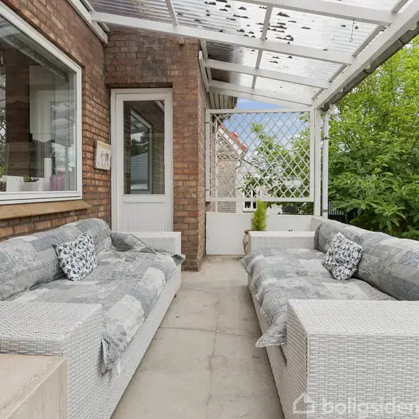 To store træer står i en have, hvor de skygger et bord med stole. I baggrunden ses en terrasse og en murstensbygning omkranset af grøn beplantning.