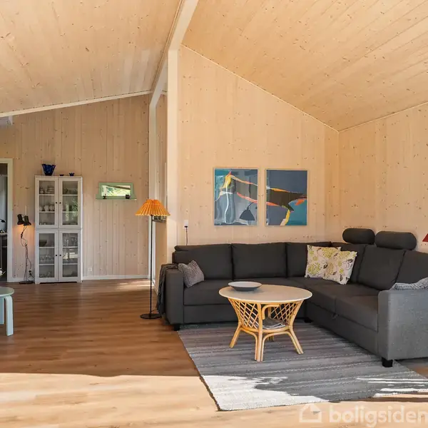 Grå hjørnesofa står i hjørnet af en stue med trævægge og højt loft; sofabord foran, farverige lamper og kunstværker omkring; spisebord og TV til venstre.
