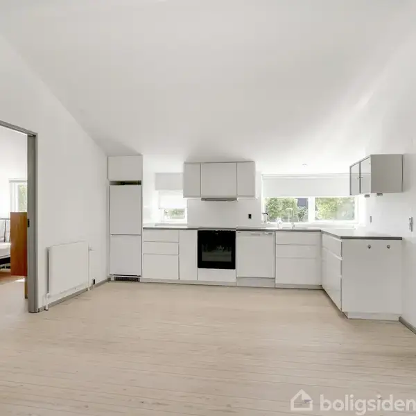 Køkken med hvide skabe og indbygget komfur. Det står i en lys, åben stue, der har en dør til et soveværelse med seng og vindue.