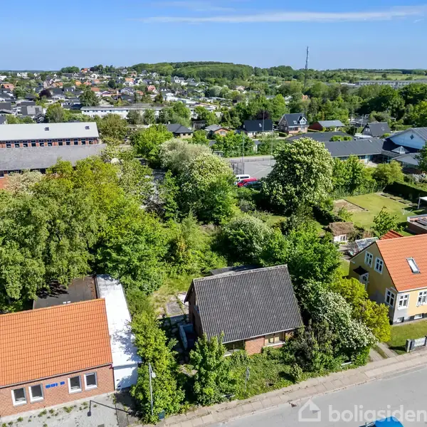 Et parcelhuskvarter med tre huse med røde og mørke tage, omgivet af grønne haver og træer, set fra oven. I baggrunden ses flere huse og en skolebygning.