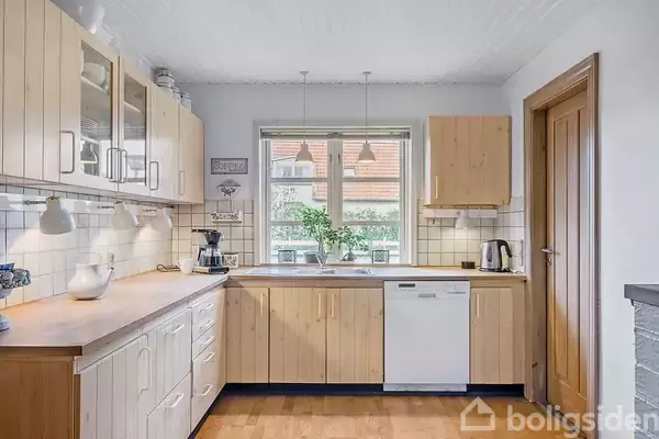 Et køkken med træskabe og hvide fliser. Der er en kaffemaskine og el-kedel på bordet. Vindue med planter ser ud til en murstensbygning.